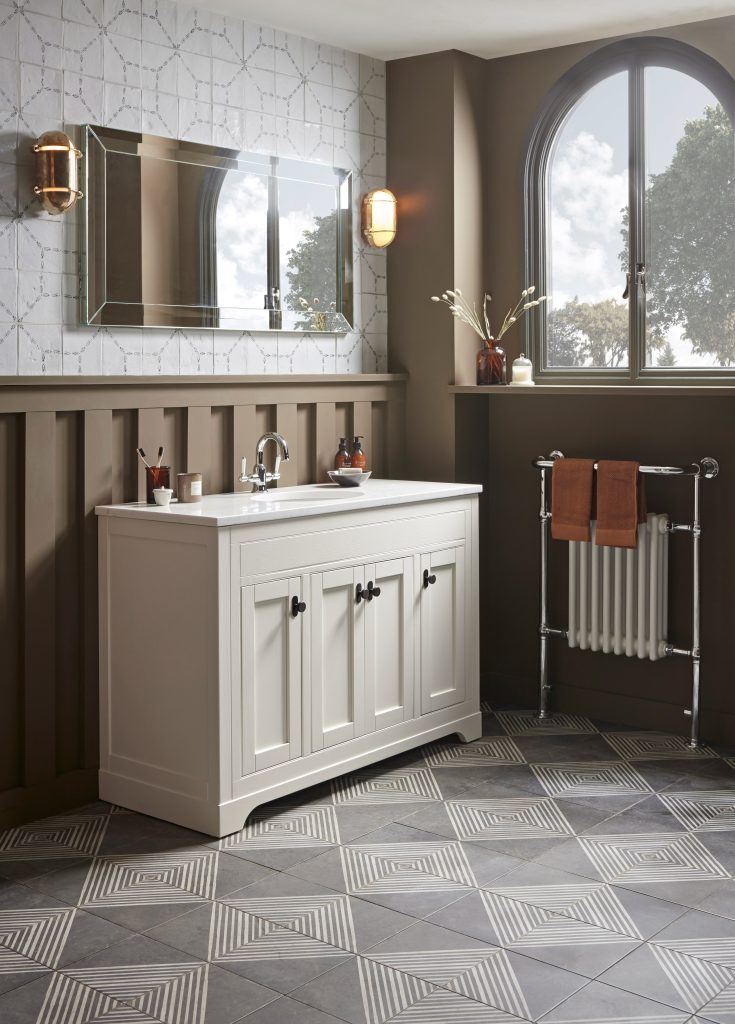 Traditional bathroom featuring a white vanity unit with chrome basin tap, bevelled wall mirror, arched window, towel radiator and grey geometric patterned floor tiles