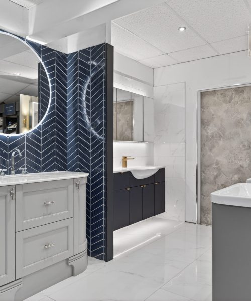 Luxury bathroom showroom featuring grey vanity units, white basins, illuminated round mirror, navy herringbone wall tiles and marble-effect flooring